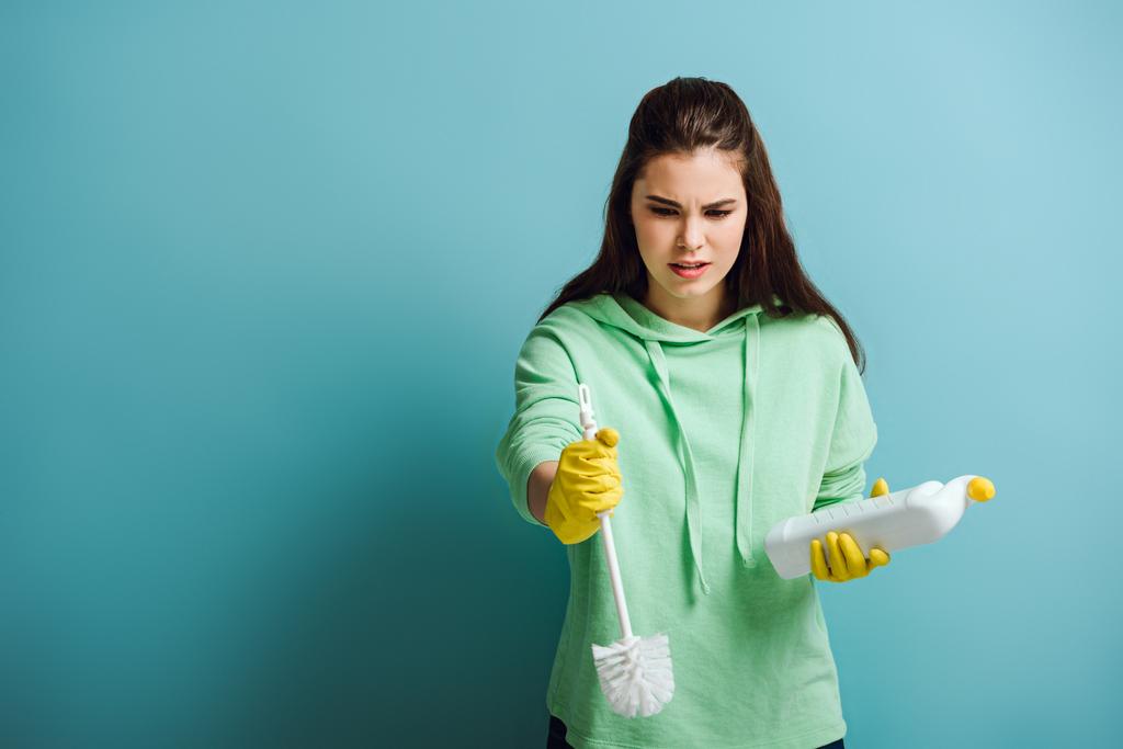 unzufriedene Hausfrau hält Toilettenreiniger und Kolben auf blauem Hintergrund - Foto, Bild