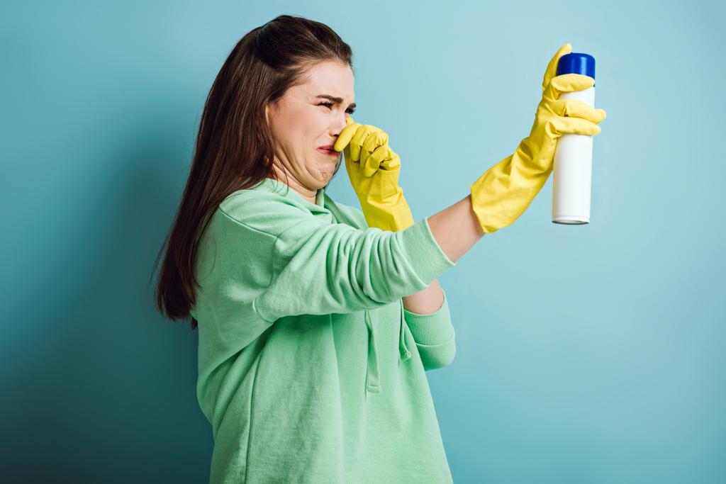 displeased housewife plugging nose with hand while spraying air freshener on blue background - Photo, Image