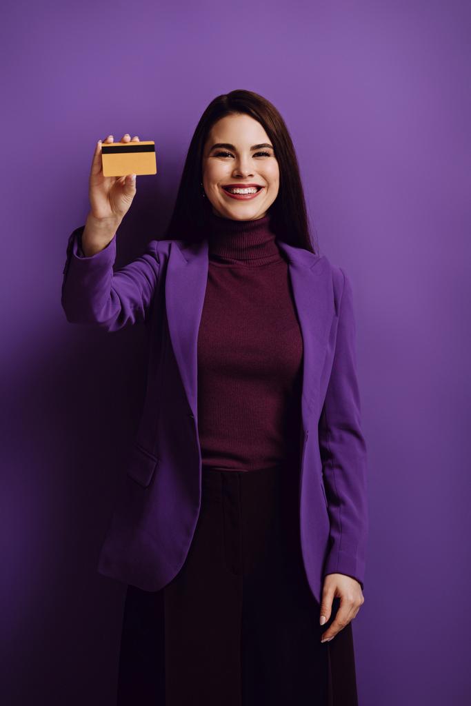 紫の背景にクレジットカードを見せながらカメラで微笑む幸せなスタイリッシュな女性 - 写真・画像