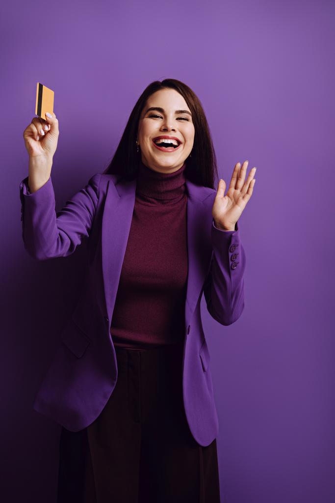 збуджена молода жінка махає рукою і сміється, тримаючи кредитну картку на фіолетовому фоні
 - Фото, зображення