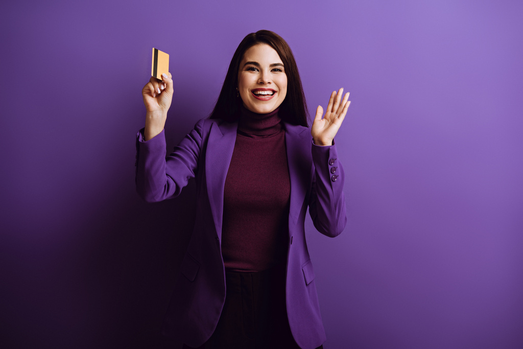 alegre joven mujer agitando la mano y sonriendo mientras sostiene la tarjeta de crédito sobre fondo púrpura
 - Foto, imagen