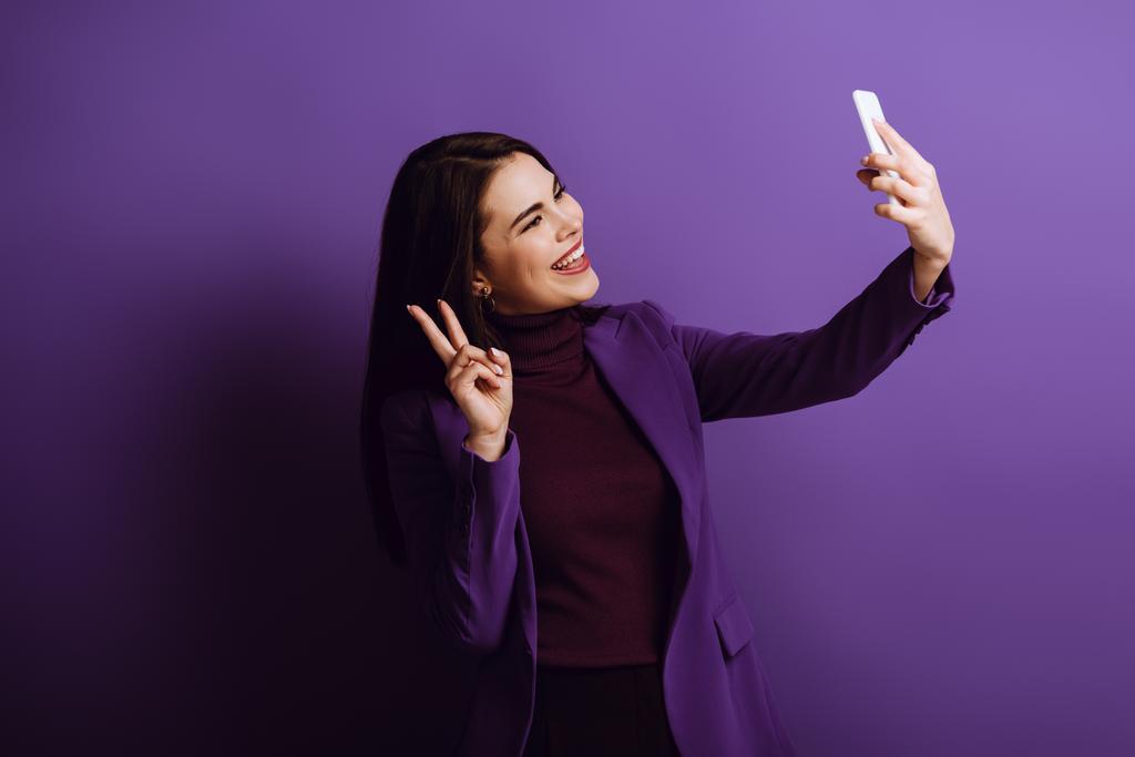 happy young woman showing victory gesture while taking selfie on purple background - Photo, Image
