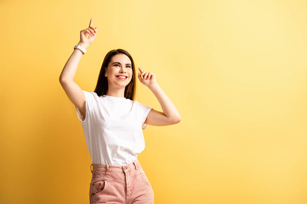 cheerful girl dancing with raised hands while looking away on yellow background - Photo, Image
