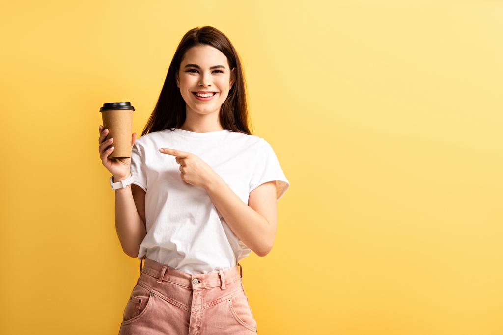 veselá dívka ukazuje prstem na kávu jít, zatímco se usmívá na kameru izolované na žluté - Fotografie, Obrázek
