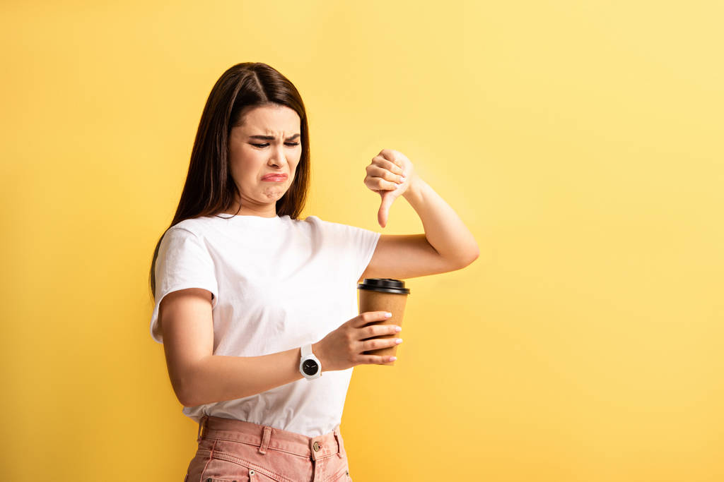 unpleased girl showing thumb down while holding coffee to go isolated on yellow
 - Фото, изображение