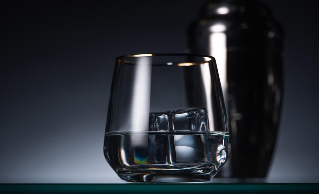 επιλεκτική εστίαση από διαφανές γυαλί με παγάκι και βότκα στο σκοτάδι με πίσω φως και αναδευτήρα - Φωτογραφία, εικόνα