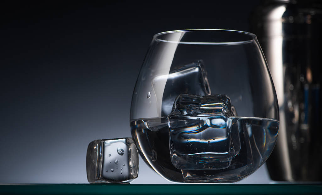 selektiver Fokus aus transparentem Glas mit Eiswürfeln und Wodka im Dunkeln mit Rücklicht und Shaker - Foto, Bild