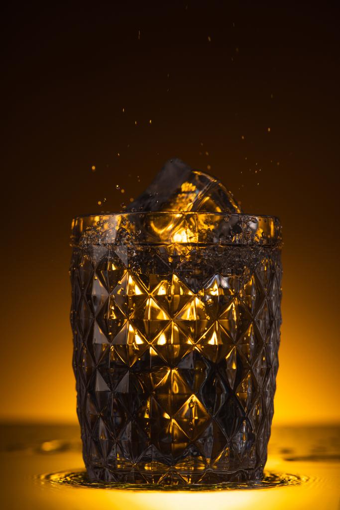 faceted glass with ice cube and splashing vodka in dark with warm back light - Photo, Image