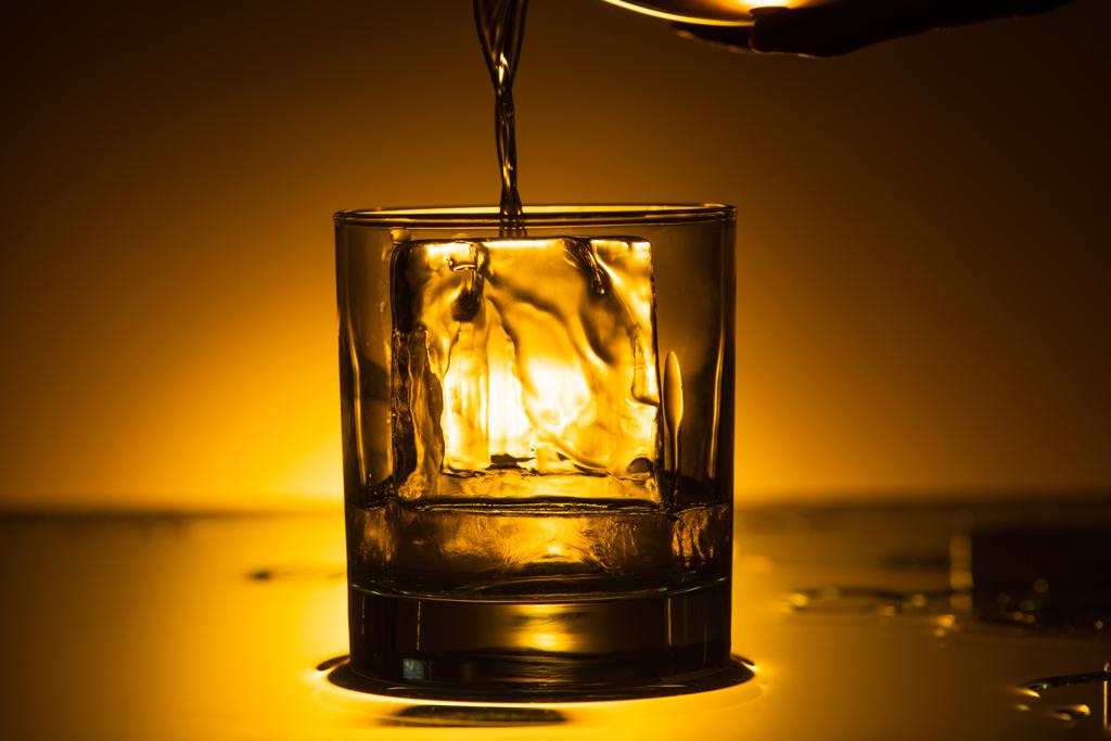 vodka derramando em vidro transparente com cubo de gelo no escuro com luz traseira quente
 - Foto, Imagem