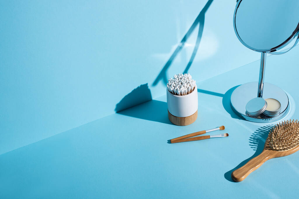 Вушні палички утримувача зубної щітки біля косметичних щіток, дзеркало з баночкою воску та щіткою для волосся на синьому фоні, концепція нульових відходів
 - Фото, зображення