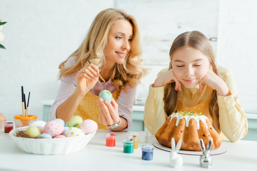 happy mother painting chicken egg near daughter, decorative rabbits and easter cake  - Photo, Image