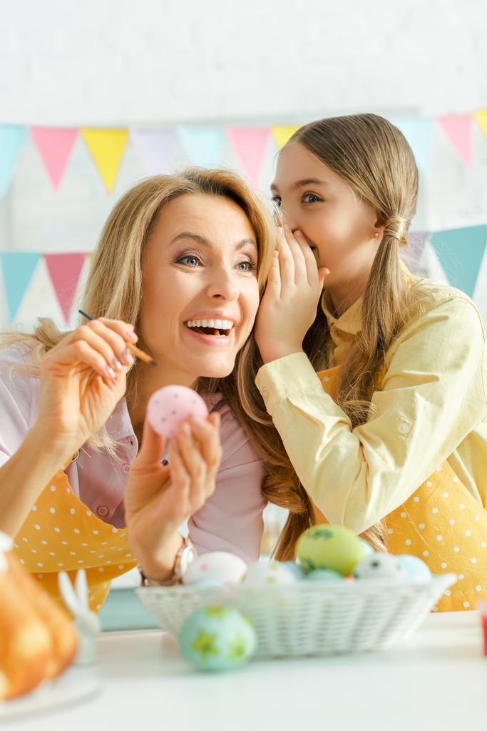 επιλεκτική εστίαση του παιδιού ψιθυρίζοντας στο αυτί της χαρούμενης μητέρας κοντά στο Πάσχα αυγά  - Φωτογραφία, εικόνα