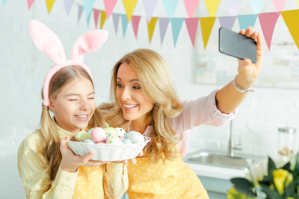 Selektiver Fokus von glücklicher Mutter und Tochter in Hasenohren beim Selfie mit bemalten Ostereiern im Korb  - Foto, Bild