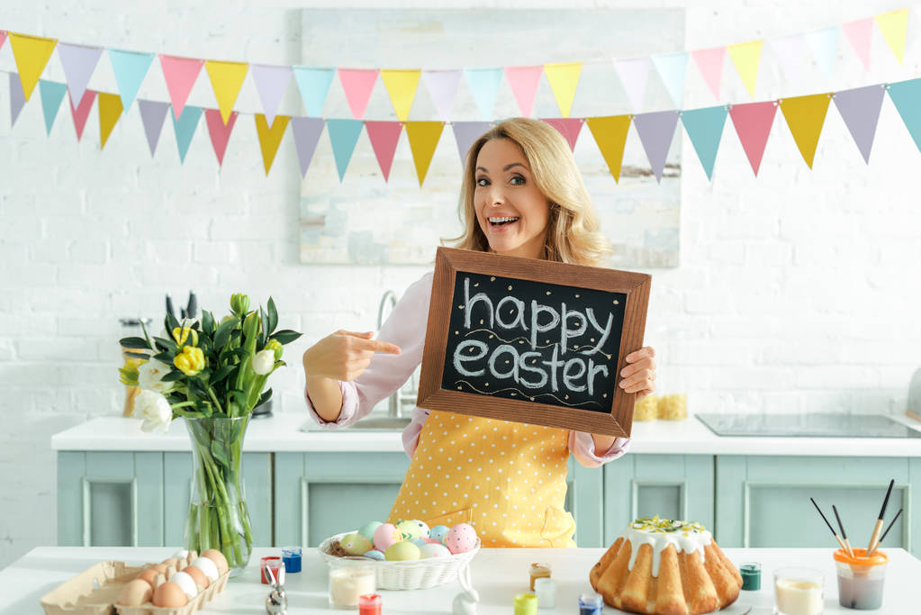 cheerful woman pointing with finger at chalkboard with happy easter lettering  - Photo, Image