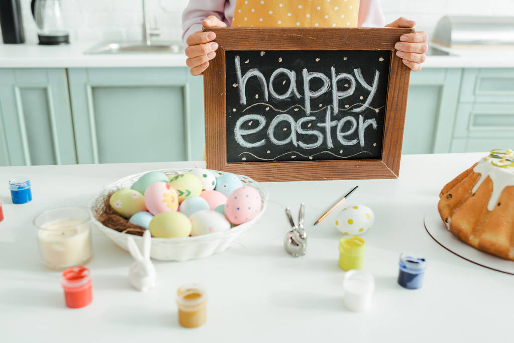 oříznutý pohled na ženu držící tabuli se šťastnými velikonočními písmeny v blízkosti dekorativních králíků a slepičích vajec  - Fotografie, Obrázek