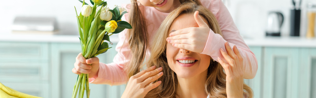 panoramatický záběr dítěte zakrývající oči šťastné matky při držení kytice tulipánů  - Fotografie, Obrázek