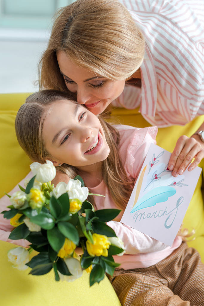 ευτυχισμένη μητέρα κρατώντας ευχετήρια κάρτα με γράμματα 8 Μαρτίου, ενώ φιλιά κόρη στο σπίτι  - Φωτογραφία, εικόνα