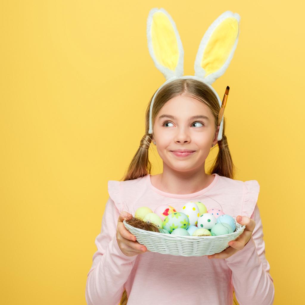 Ευτυχισμένο παιδί με αυτιά λαγού κρατώντας αυγά Πάσχα στο καλάθι και κοιτάζοντας μακριά απομονώνονται σε κίτρινο  - Φωτογραφία, εικόνα