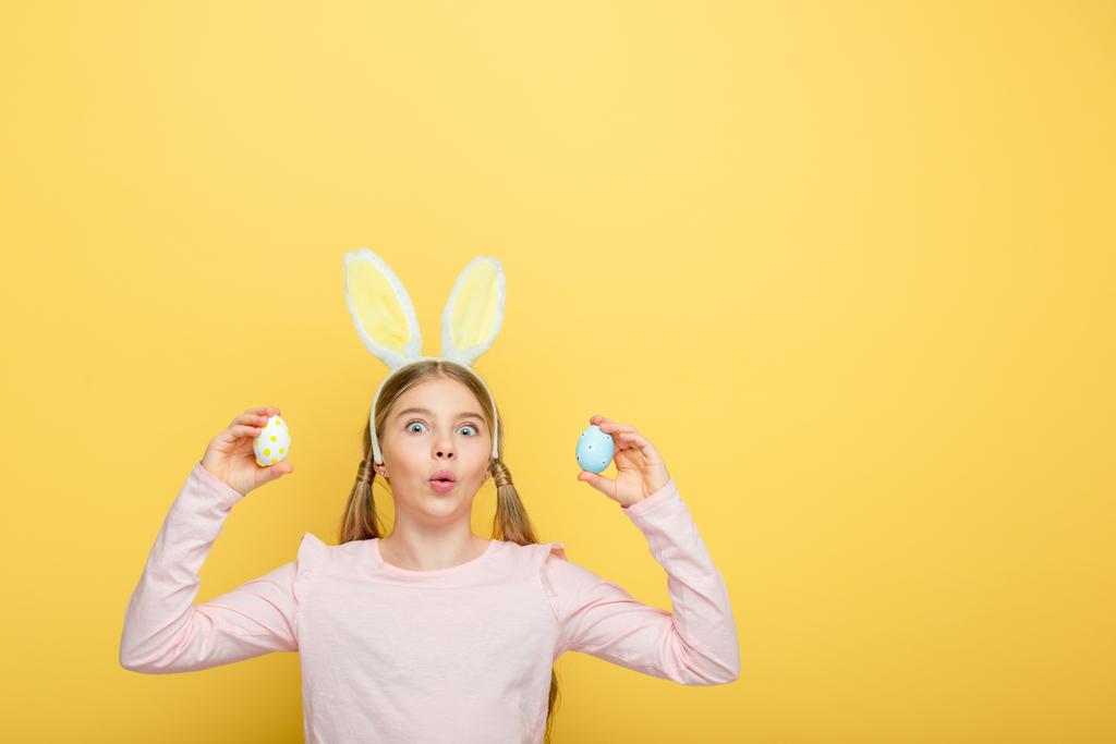 niño sorprendido con orejas de conejo sosteniendo huevos de Pascua pintados aislados en amarillo
  - Foto, Imagen