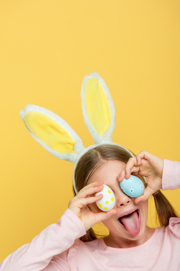 enfant avec des oreilles de lapin collant la langue et couvrant les yeux avec des œufs de Pâques isolés sur jaune
  - Photo, image