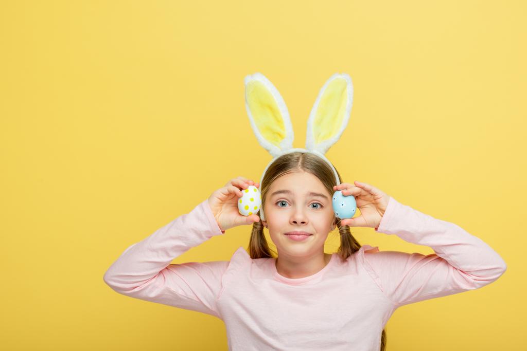 cute child with bunny ears holding painted easter eggs isolated on yellow  - Photo, Image