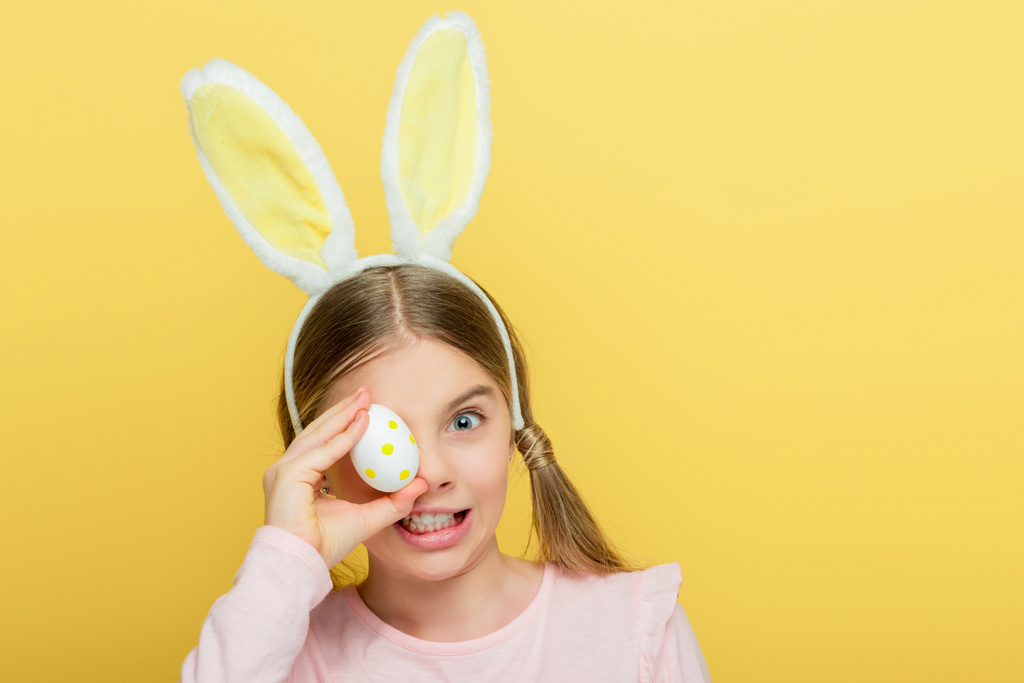 emocjonalne dziecko z króliczymi uszami zakrywającymi oko jajkami wielkanocnymi wyizolowanymi na żółto  - Zdjęcie, obraz
