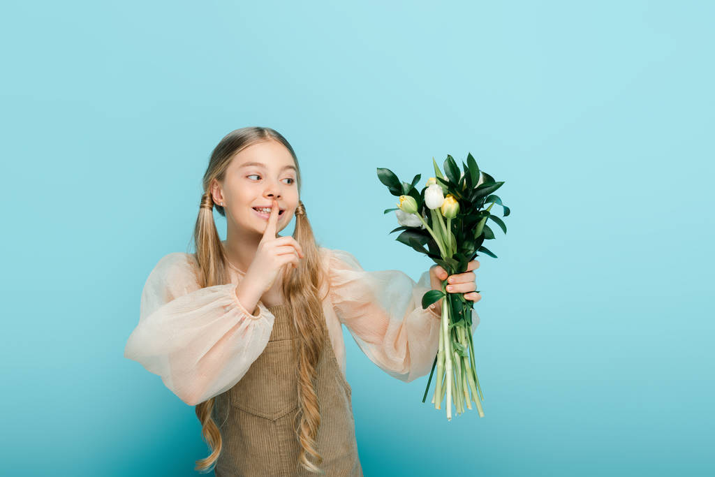 happy kid looking at bouquet of tulips and showing hush sign isolated on blue  - Photo, Image