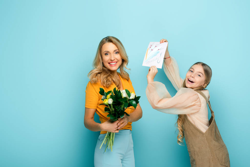 glückliche Tochter hält Grußkarte mit 8 März Schriftzug neben Mutter mit Tulpen auf blau - Foto, Bild
