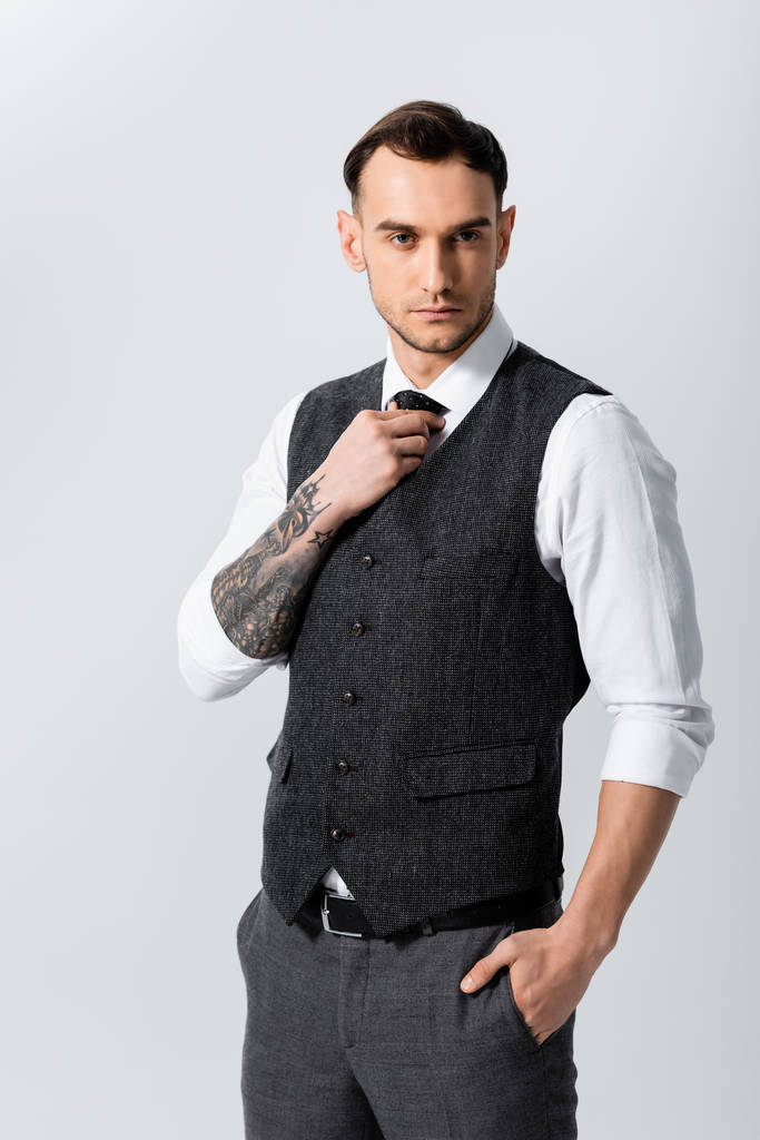 guapo tatuado elegante novio con mano en bolsillo ajuste corbata aislado en gris
 - Foto, Imagen