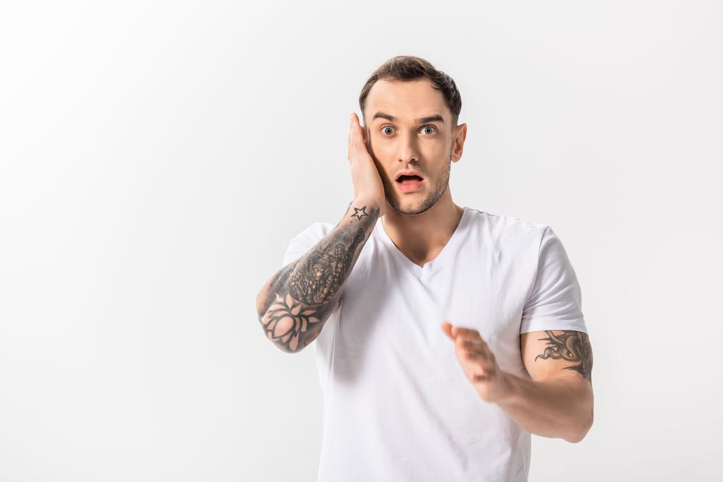 σοκαρισμένος όμορφος νεαρός άνδρας με τατουάζ αγγίζοντας πρόσωπο απομονωμένο σε λευκό - Φωτογραφία, εικόνα