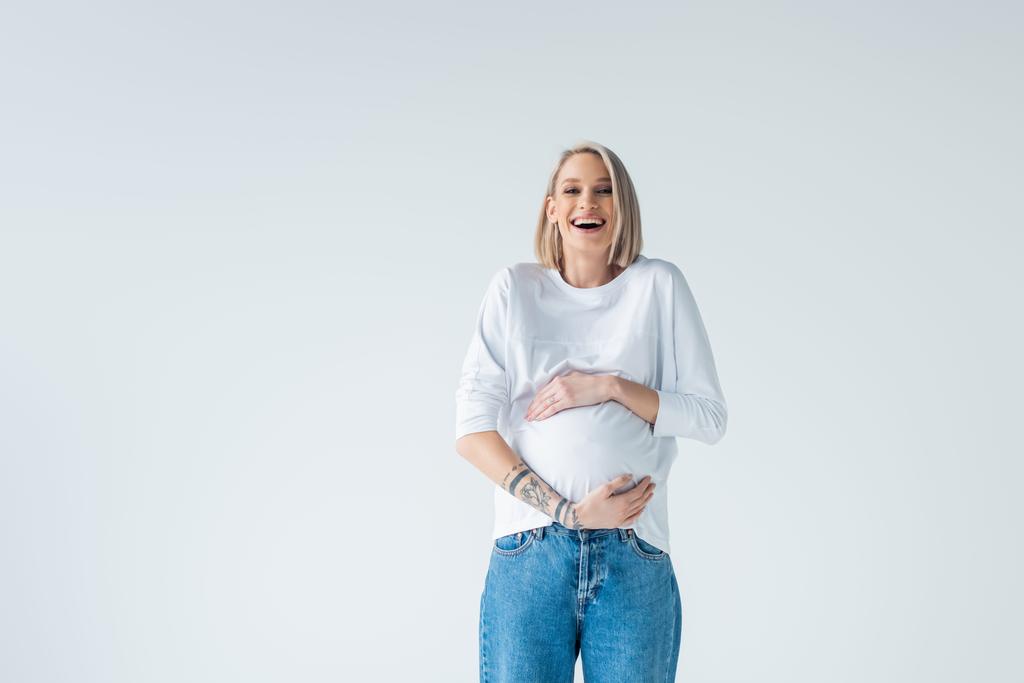 šťastná mladá tetovaná těhotná žena dotýkající se břicha izolované na šedé - Fotografie, Obrázek
