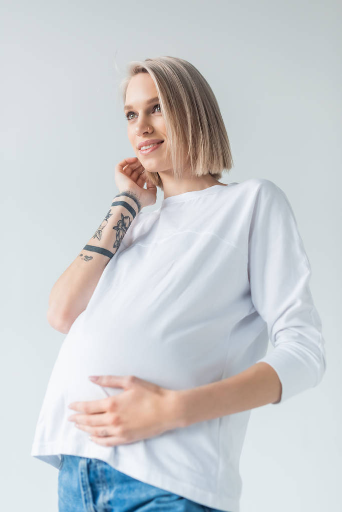 hymyilevä nuori tatuoitu raskaana oleva nainen koskettaa vatsa eristetty harmaa
 - Valokuva, kuva