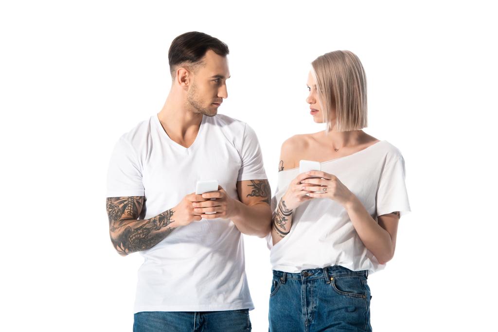 νεαρό ζευγάρι με τατουάζ που χρησιμοποιεί smartphones και κοιτάζει ο ένας τον άλλον απομονωμένο στα λευκά - Φωτογραφία, εικόνα