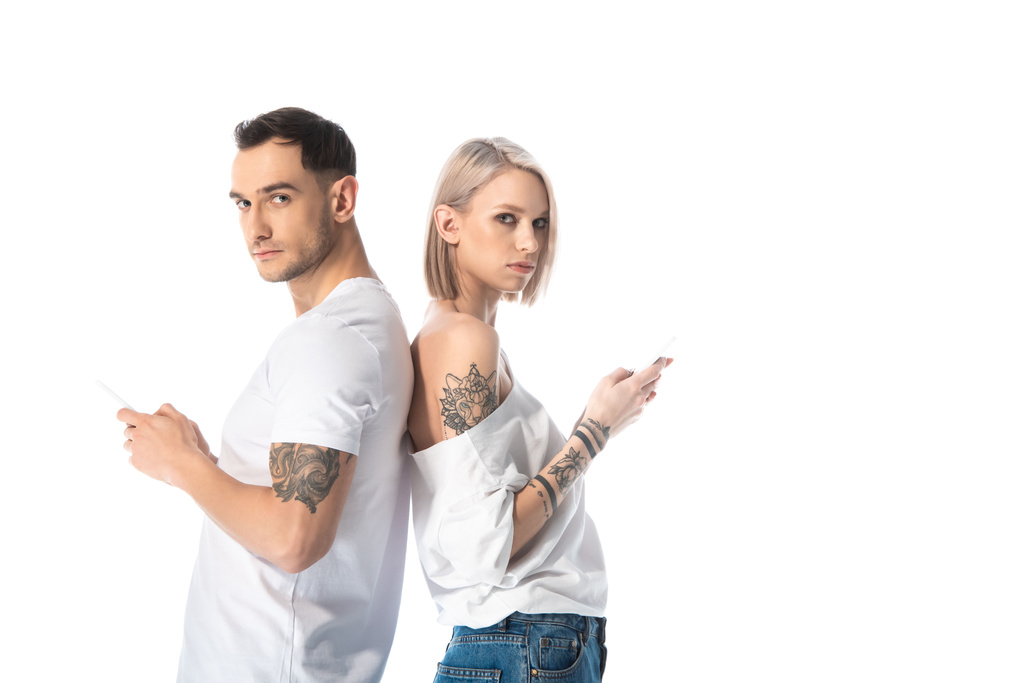 joven pareja tatuada utilizando teléfonos inteligentes, mientras que de pie espalda con espalda aislado en blanco
 - Foto, imagen