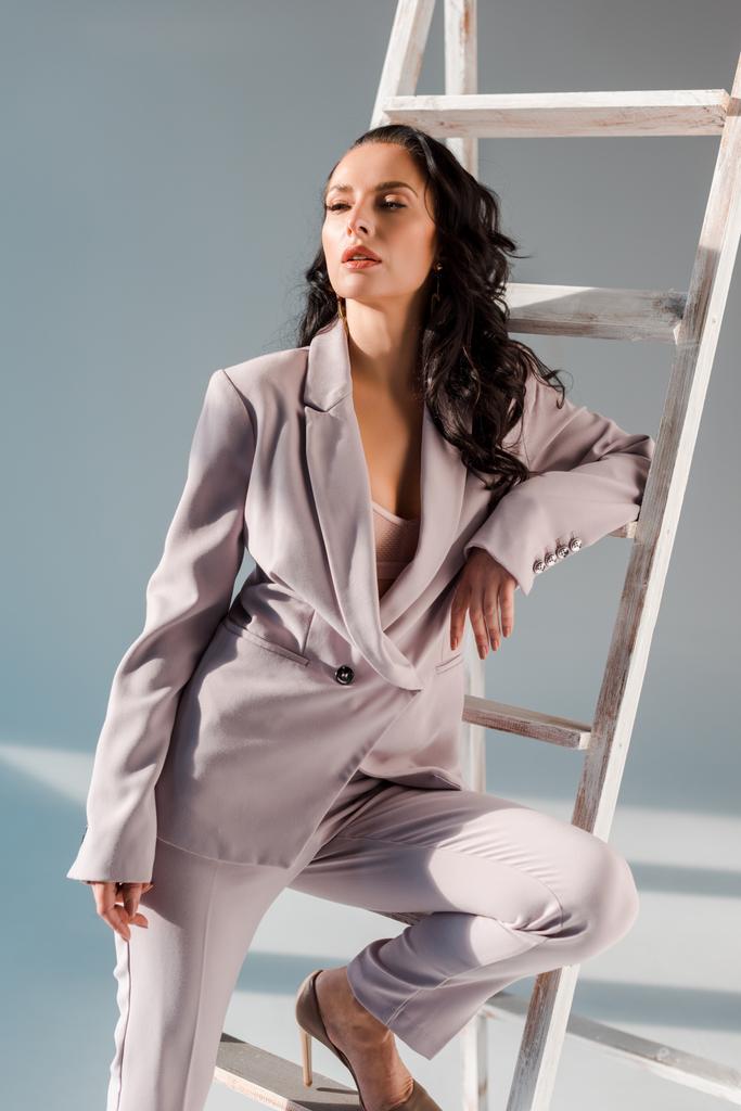 stylische Frau im Anzug in der Nähe der Leiter, die vor grauem Hintergrund wegschaut  - Foto, Bild