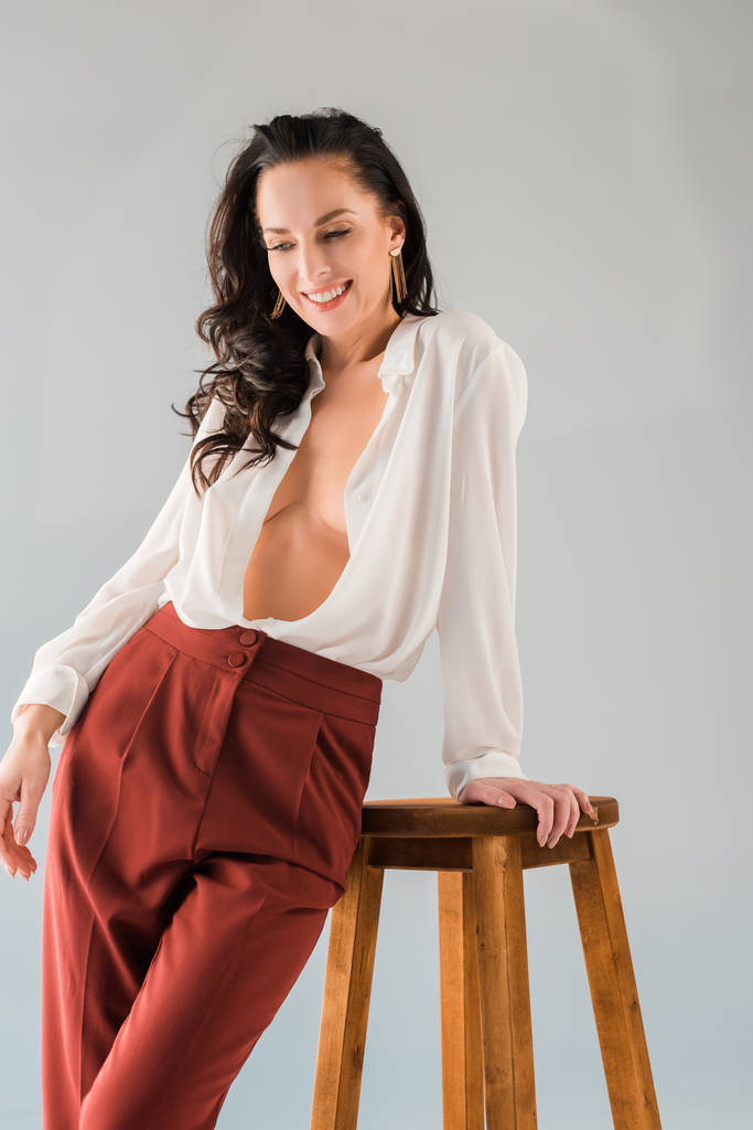 smiling and sexy woman in shirt and trousers standing near stool isolated on grey  - Photo, Image