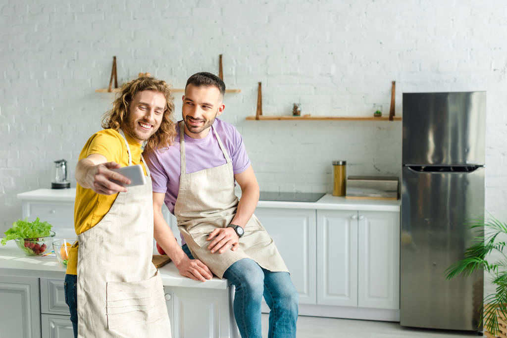 selectieve focus van gelukkige homoseksuele mannen in schorten selfie in de keuken  - Foto, afbeelding