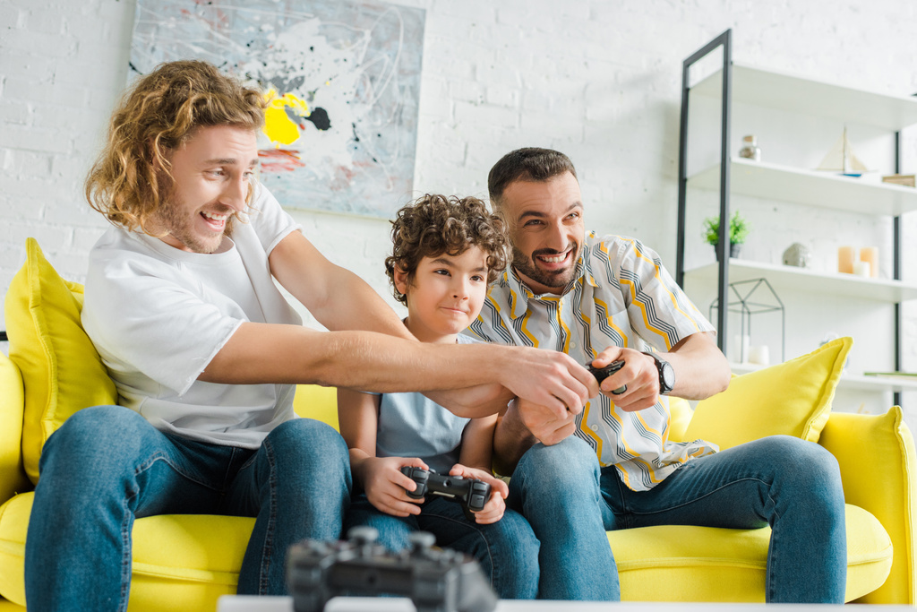 KYIV, UCRAINA - 28 GENNAIO 2020: focus selettivo della coppia omosessuale felice che gioca al videogioco con il figlio di razza mista
  - Foto, immagini