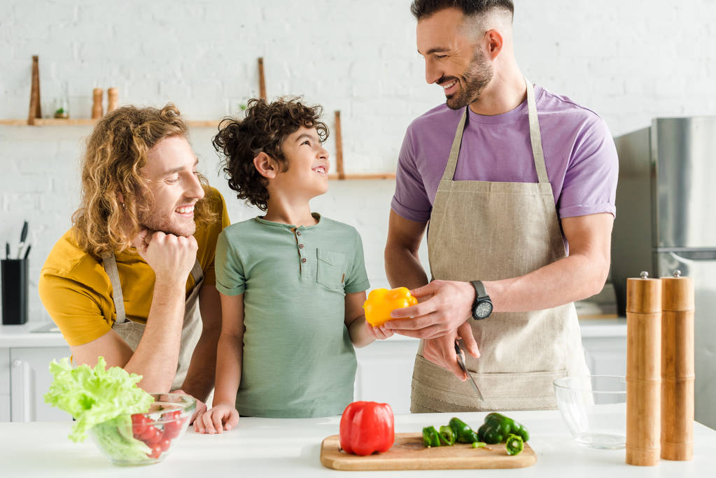 кудрявый ребенок смешанной расы рядом с счастливыми гомосексуальными родителями на кухне
  - Фото, изображение
