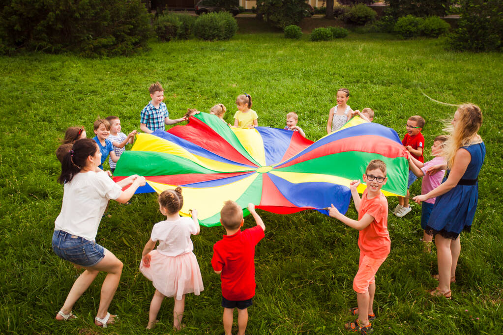 Lapset leikkivät sateenkaaren laskuvarjolla puistossa.
 - Valokuva, kuva
