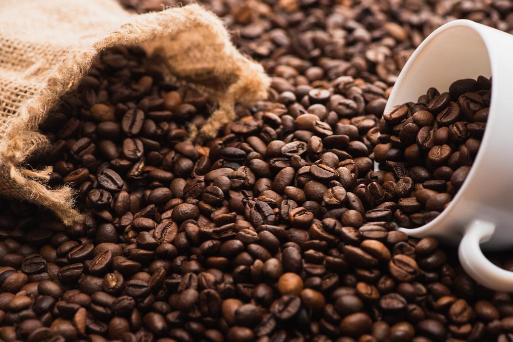 селективный фокус свежих жареных кофейных зерен и белая чашка возле мешка
 - Фото, изображение