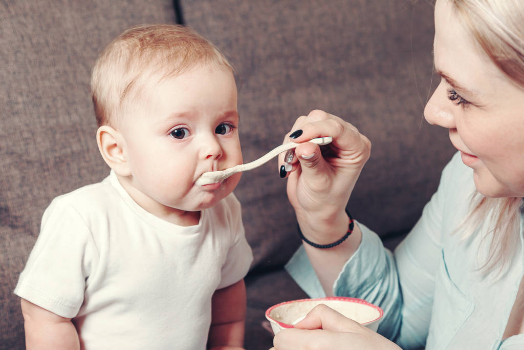 Een gelukkige moeder of oppas voedt een kleine baby met een lepel voedsel. Het gebrek aan onafhankelijkheid van een klein kind - Foto, afbeelding