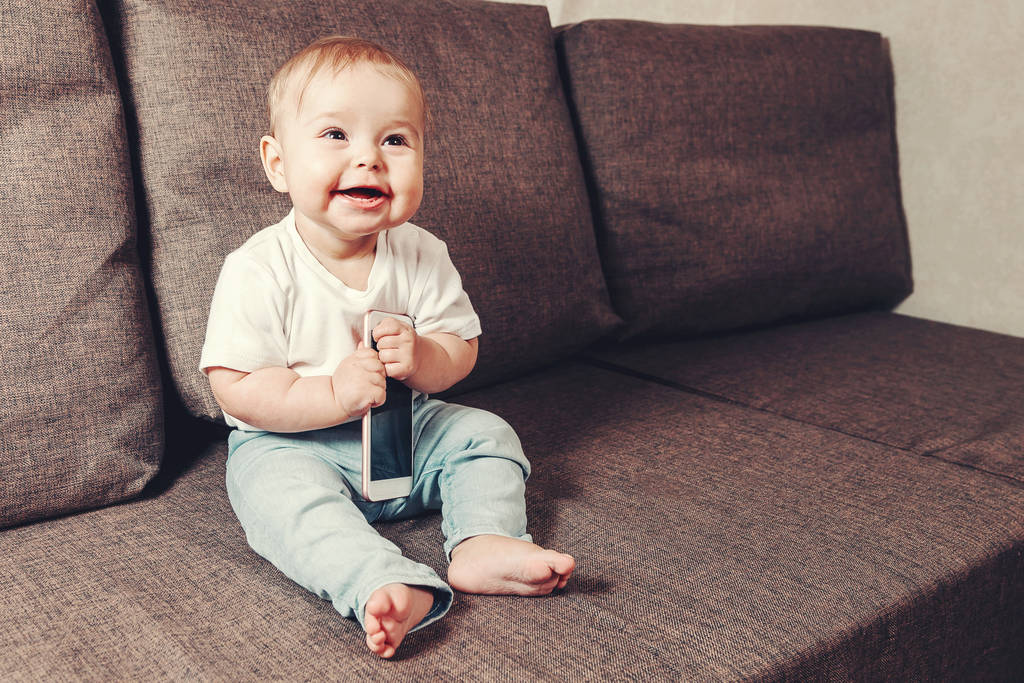 Ребенок узнаёт мир, современные детские игрушки. Маленький улыбающийся ребенок сидит на диване с телефоном в руке. копировальное пространство
 - Фото, изображение