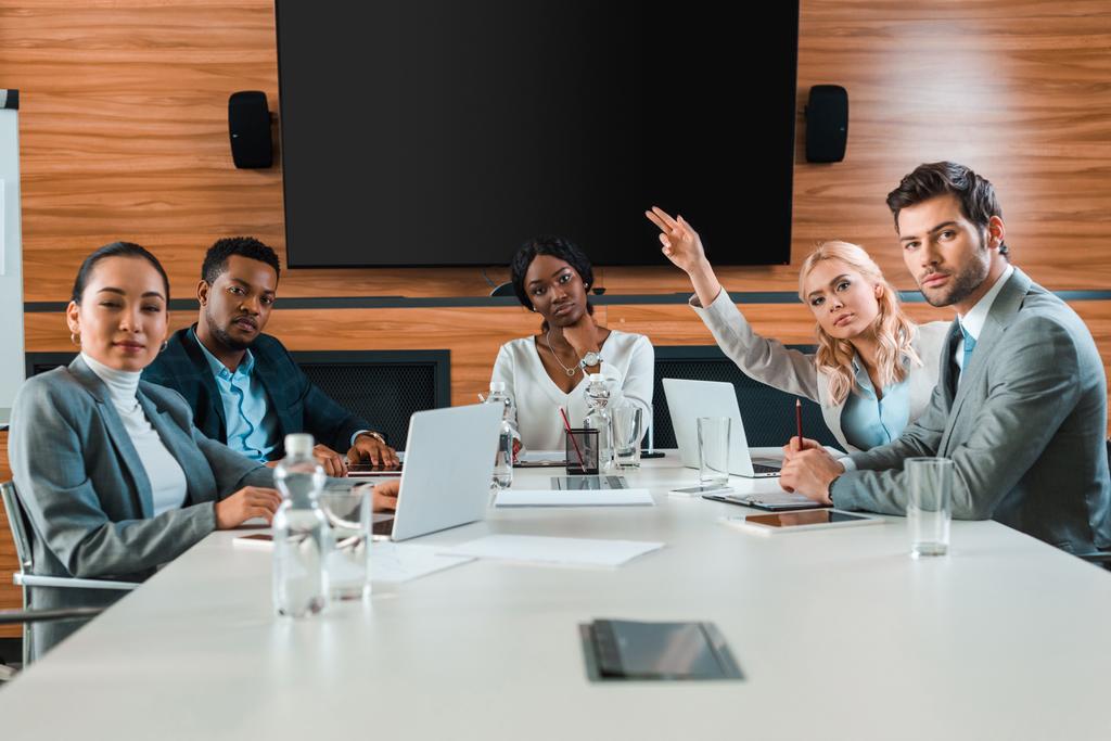 jonge zakenvrouw hand opsteken terwijl zitten in conferentiezaal in de buurt van multiculturele collega 's op zoek naar camera - Foto, afbeelding