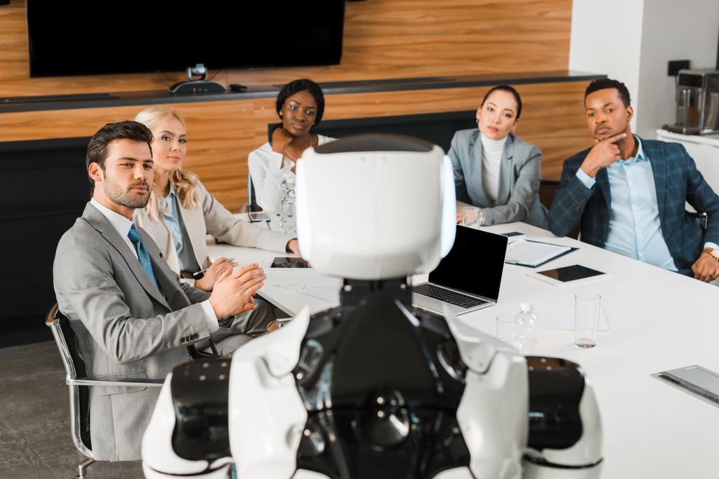 selective focus of young multicultural businesspeople looking at robot while sitting in conference hall - Photo, Image