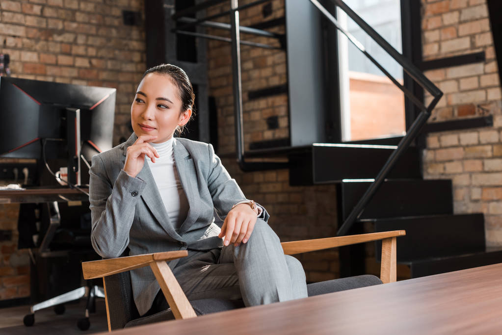 atractivo asiático mujer de negocios sentado en sillón y tocando barbilla mientras mira lejos
   - Foto, Imagen