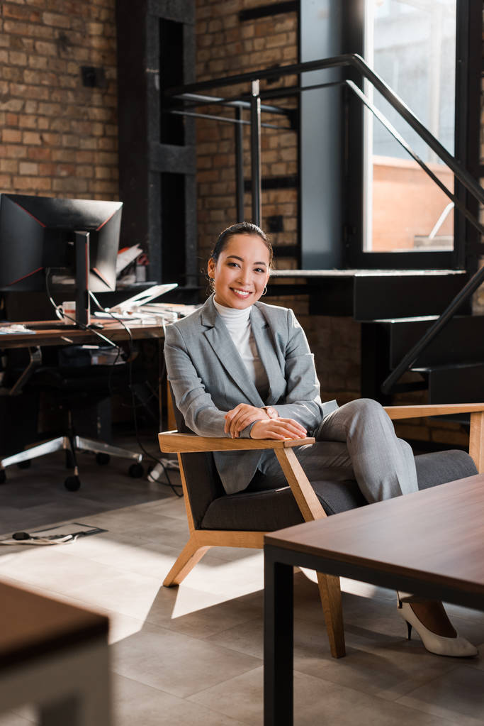 избирательный фокус привлекательной азиатской предпринимательницы, сидящей в кресле и улыбающейся в камеру
 - Фото, изображение