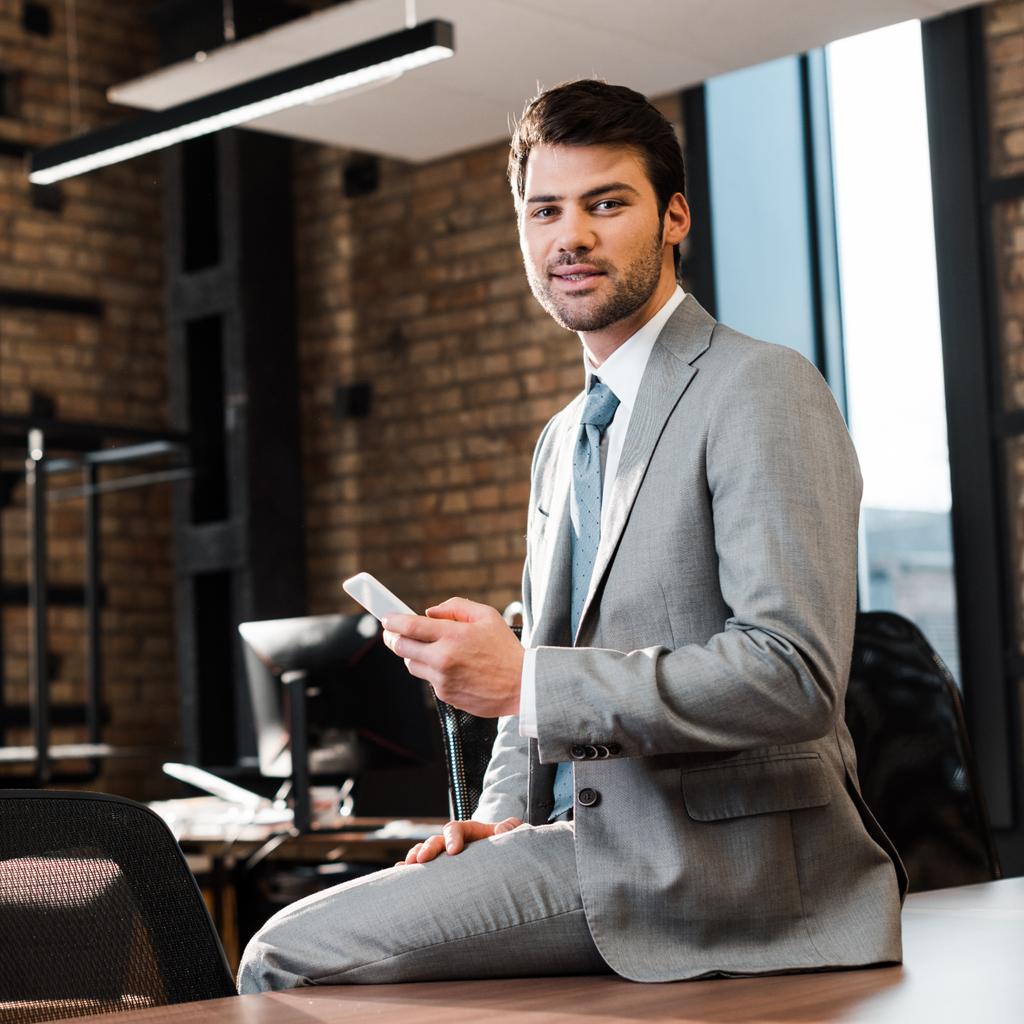 pohledný, usměvavý obchodník sedící na kancelářském stole, držící smartphone a dívající se do kamery - Fotografie, Obrázek