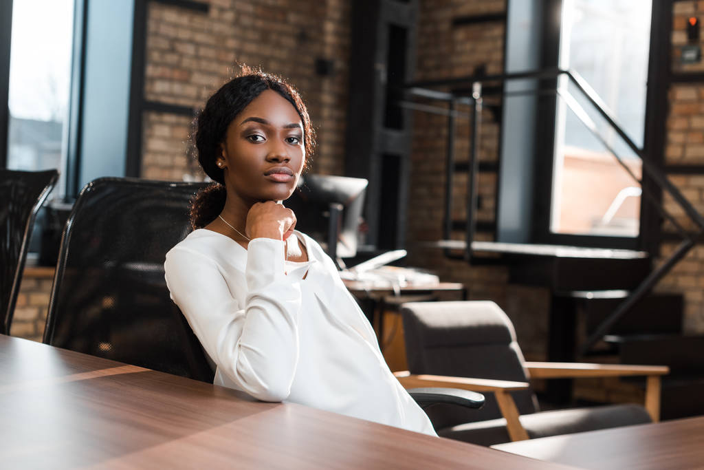 thoughtful, pregnant african american businesswoman sitting at office desk and looking at camera - Photo, Image