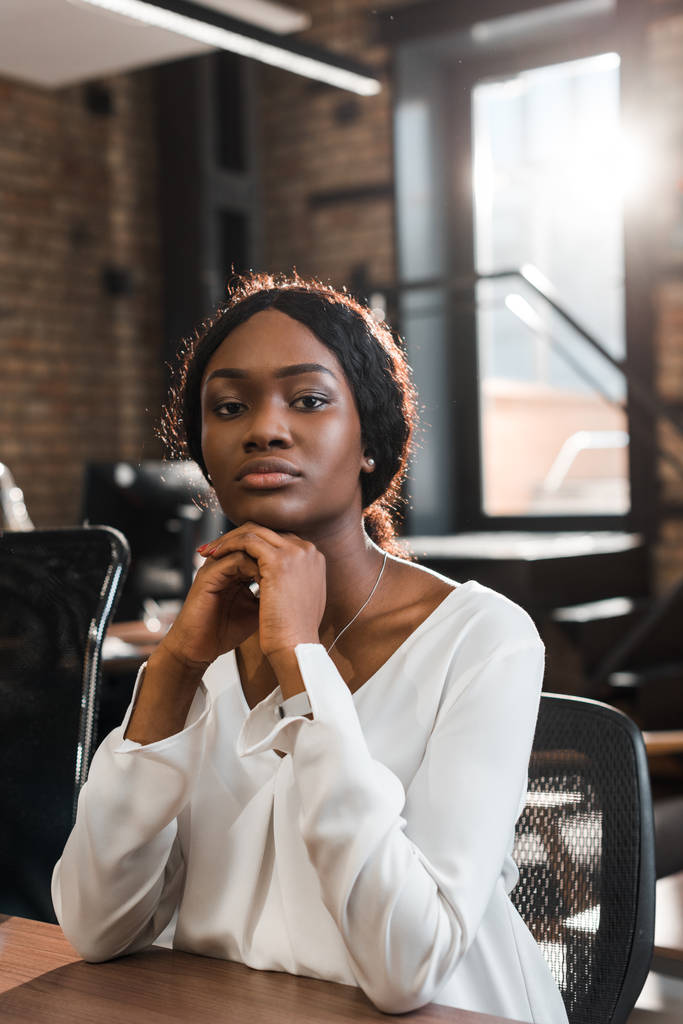 pensive, attractive african american businesswoman sitting in office and looking at camera - Photo, Image
